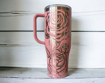 May include: A pink stainless steel travel mug with a handle. The mug has a gold floral design with roses.