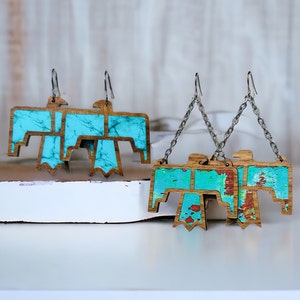 May include: Wooden earrings in the shape of a thunderbird, featuring a turquoise and brown color scheme. The earrings have a distressed, painted look and are suspended from silver-colored ear wires.