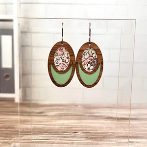 May include: A pair of oval-shaped wooden earrings with a floral pattern inside a green oval cutout. The earrings have silver hooks.
