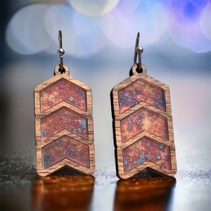 Puede incluir: Un par de pendientes de madera con un patrón de chevron. Los pendientes están pintados con una pintura de color óxido y tienen un acabado de purpurina azul metálico.