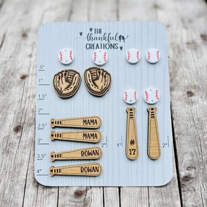 Könnte beinhalten: Eine Holztafel mit verschiedenen Baseball-Ohrringen. Die Ohrringe sind aus Holz gefertigt und zeigen Baseballschläger, Baseballhandschuhe und Baseballs. Die Tafel enthält auch ein Lineal zum Messen der Größe der Ohrringe. Der Text "The Thankful Creations" ist auf der Tafel aufgedruckt.