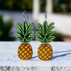 May include: A pair of wooden pineapple earrings with green tops and yellow bottoms. The earrings are hanging from silver hooks.