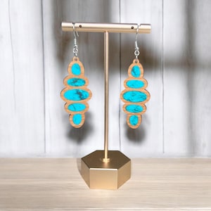 May include: A pair of turquoise and wood earrings with a geometric design. The earrings are hanging from a gold stand.