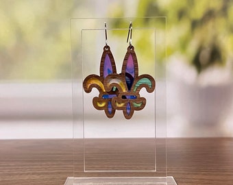 May include: A pair of colorful wooden earrings shaped like fleur-de-lis. The earrings are made of layered wood with a purple, blue, green, and yellow color scheme. The earrings are hanging from silver hooks.
