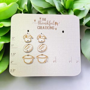 May include: A light-colored wooden display board with laser-cut food-themed charms. The charms include jars of jam, bread, and a cooking pot. The text "THE thankful CREATIONS" is at the top. Measurements are in inches.