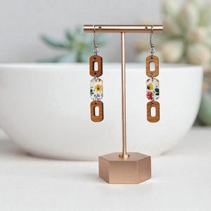May include: A pair of dangle earrings displayed on a gold stand. Each earring features three wooden rectangular shapes, with a floral design in the middle. The earrings have silver hooks and are set against a soft, blurred background.