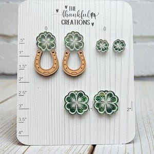Op de afbeelding: Een set oorbellen voor St. Patrick's Day op een witte kaart. De oorbellen hebben groene en witte klaverontwerpen, sommige met hoefijzers. De kaart toont de tekst "THE thankful CREATIONS". De afmetingen zijn in inches.