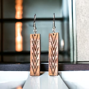 May include: A pair of wooden earrings with a geometric chevron pattern. The earrings are brown with white accents and have silver hooks.