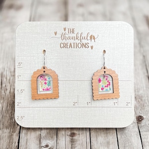 May include: A pair of wooden earrings with a floral pattern inside the cutout. The earrings are 1 inch wide and 1.5 inches tall. The earrings are on a display card with the text "THE thankful CREATIONS".