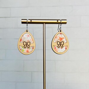 May include: A pair of wooden earrings shaped like Easter eggs with a floral pattern and a cutout bow. The earrings are displayed on a gold stand.