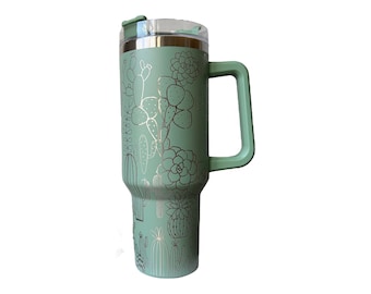 May include: A green stainless steel travel mug with a black handle and a cactus and succulent design. The mug has a lid with a flip top.