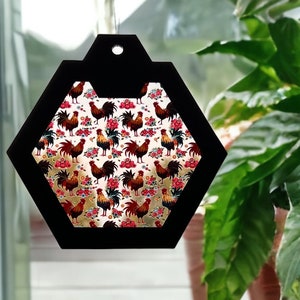 Pode incluir: Um ornamento hexagonal preto com fundo branco e um padrão repetido de galos vermelhos, marrons e amarelos. O ornamento tem um furo na parte superior para pendurar.