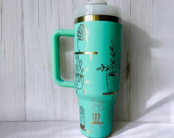 May include: A turquoise travel mug with a gold lid and handle. The mug has a gold design of potted plants and succulents.