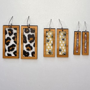 May include: Six pairs of wooden earrings with different patterns. The patterns include leopard print, bees and honeycomb, and a pink and white striped pattern.