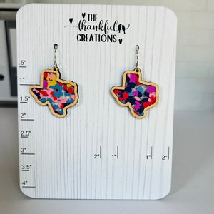 May include: A pair of dangle earrings featuring a Texas state outline with a colorful floral pattern. The earrings are displayed on a white wooden board with a ruler for size reference. The board also includes the text "THE thankful CREATIONS".