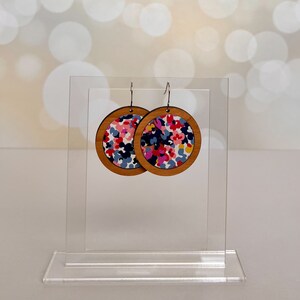May include: A pair of round wooden earrings with a colorful floral pattern. The earrings are displayed on a clear acrylic stand.