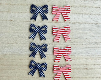 May include: Eight wooden bow-shaped ornaments. Four are navy blue with white star patterns, and four are red with white stripes. The ornaments are arranged on a light wood-grain surface.
