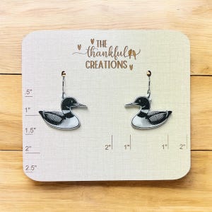 May include: A pair of earrings with a black and white loon design. The earrings are displayed on a light-coloured card with the text "THE thankful CREATIONS". The card has inch measurements along the side.