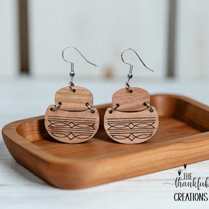May include: Wooden earrings with a geometric design. The earrings are made of light-colored wood and feature a carved pattern of lines and diamonds. They hang from silver-colored ear wires. The earrings are displayed on a wooden tray.