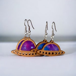 Puede incluir: Un par de pendientes de madera con forma de platillos volantes con estampado de galaxia. Los pendientes son de color marrón claro con ganchos plateados.
