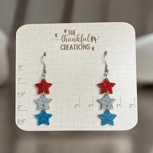May include: Earrings featuring three star-shaped charms in red, silver glitter, and blue. The earrings are displayed on a cream-colored card. The top star is red, the middle one silver glitter, and the bottom one blue. The card has the text "THE thankful CREATIONS".