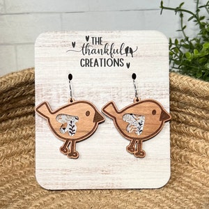 May include: A pair of wooden bird earrings with a floral fabric insert. The earrings are on a white wooden display with the text "THE thankful CREATIONS" on it.