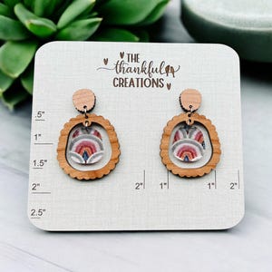 May include: A pair of wooden earrings with a scalloped edge. The earrings feature a round, clear, resin insert with a colorful, geometric pattern. The earrings are displayed on a wooden card with a ruler for size reference. The card also features the text "THE thankful CREATIONS".