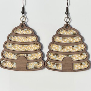 May include: A pair of wooden earrings shaped like beehives with a honeycomb pattern and gold glitter accents.