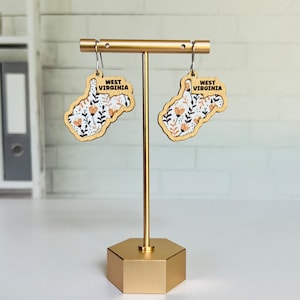 May include: A pair of wooden earrings shaped like the state of West Virginia, with a floral pattern and the words "West Virginia" inscribed on each earring. The earrings are displayed on a gold stand.