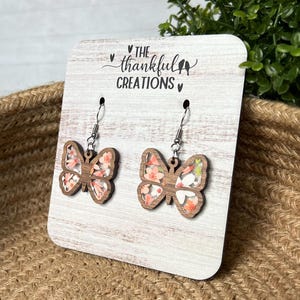 May include: A pair of wooden butterfly earrings with a floral print. The earrings are on a white wooden display card with the text "The Thankful Creations".