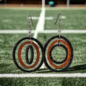 May include: A pair of black and orange glitter hoop earrings with three concentric circles. The earrings have silver hooks.