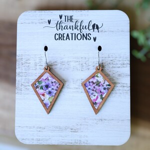 May include: A pair of diamond-shaped earrings with a floral print. The earrings are made of wood and have silver hooks. The earrings are displayed on a white wooden stand with the text "The Thankful Creations" printed on it.
