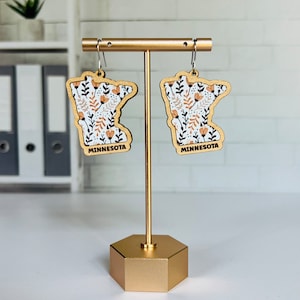 May include: A pair of wooden earrings shaped like the state of Minnesota, with a floral pattern in white, black, and orange. The earrings are displayed on a gold stand. The word "MINNESOTA" is printed on each earring.