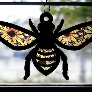 Puede incluir: Silueta negra de una abeja con alas con estampado de girasol. La abeja está colgada de una ventana.