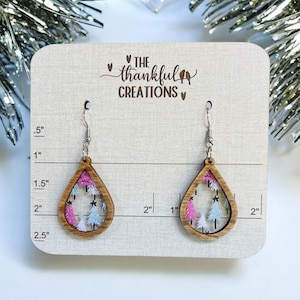May include: A pair of teardrop-shaped wooden earrings with a pink and white glitter design inside. The earrings are on a white card with a ruler and the text "The Thankful Creations".