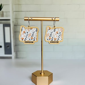 May include: A pair of wooden earrings shaped like the state of Oregon, with a floral pattern in shades of orange, white, and brown. The earrings are hanging from a gold stand and have the word "OREGON" printed on the bottom.