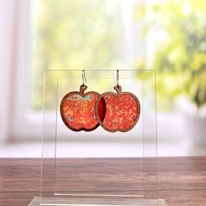 May include: A pair of apple-shaped earrings with a red and orange marbled design. The earrings are made of wood and have silver hooks.