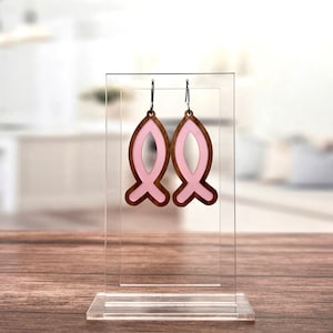 May include: A pair of pink and brown wooden earrings shaped like fish. The earrings are displayed on a clear acrylic stand.