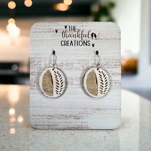 May include: A pair of wooden earrings with a leaf design. The earrings are on a white wooden display stand with the text "The Thankful Creations" on it.