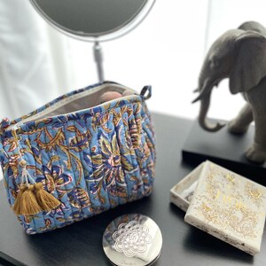 May include: A blue quilted cosmetic bag with a floral pattern, open to reveal makeup brushes. A silver compact and a small Dior box sit nearby. A decorative elephant statue and a mirror are in the background.