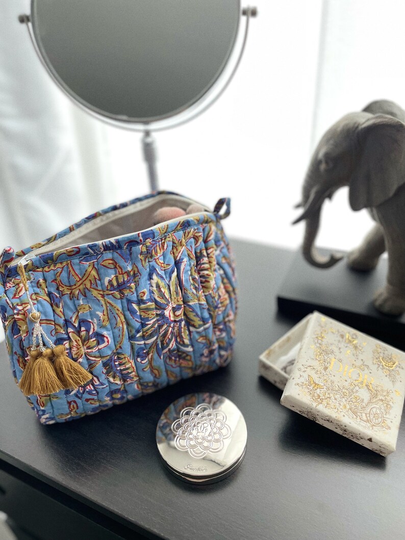 May include: A blue quilted cosmetic bag with a floral pattern, open to reveal makeup brushes. A silver compact and a small Dior box sit nearby. A decorative elephant statue and a mirror are in the background.