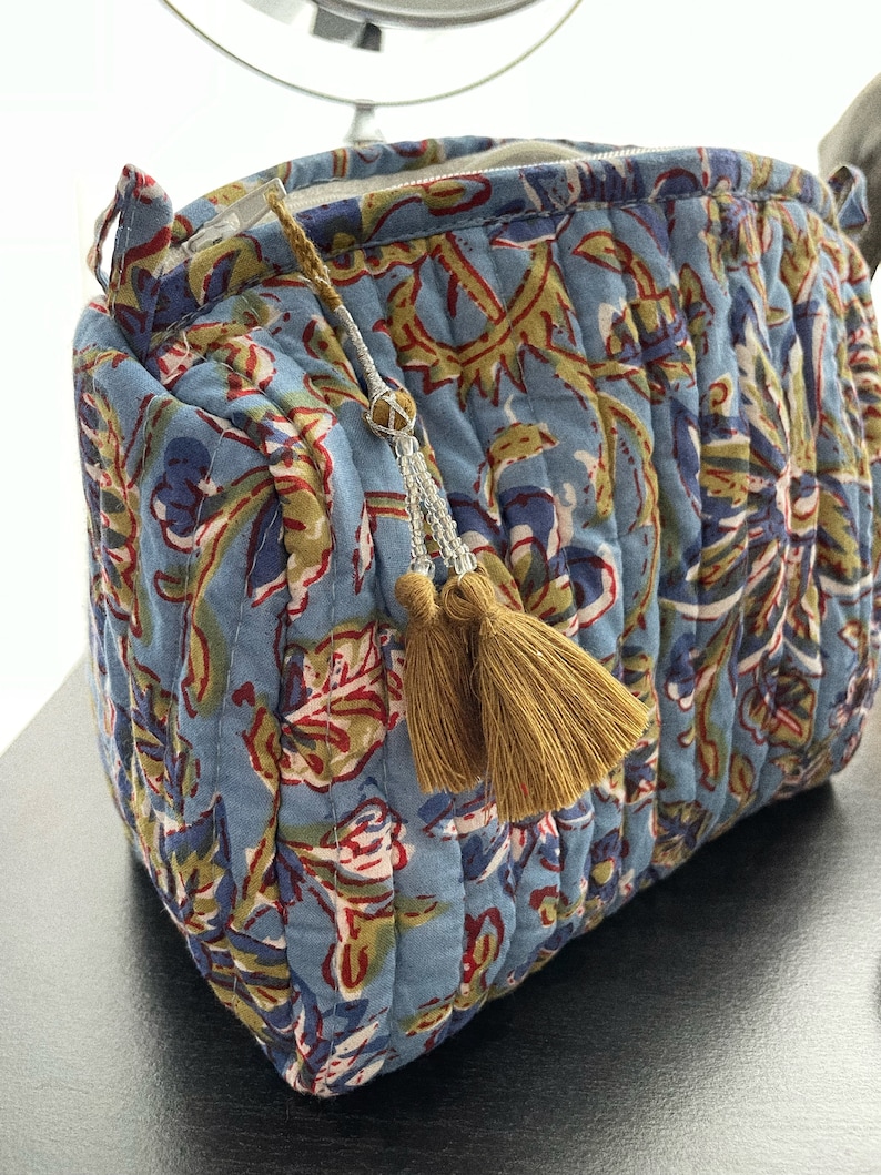 May include: A quilted cosmetic bag in a light blue fabric with a floral pattern in red, green, and navy. The bag has a gold zipper and a tassel with gold beads and two gold tassels.