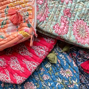 May include: Close-up of several quilted cosmetic bags in various colors. The bags feature floral patterns in shades of pink, orange, blue, and red. Each bag has a tassel zipper pull.