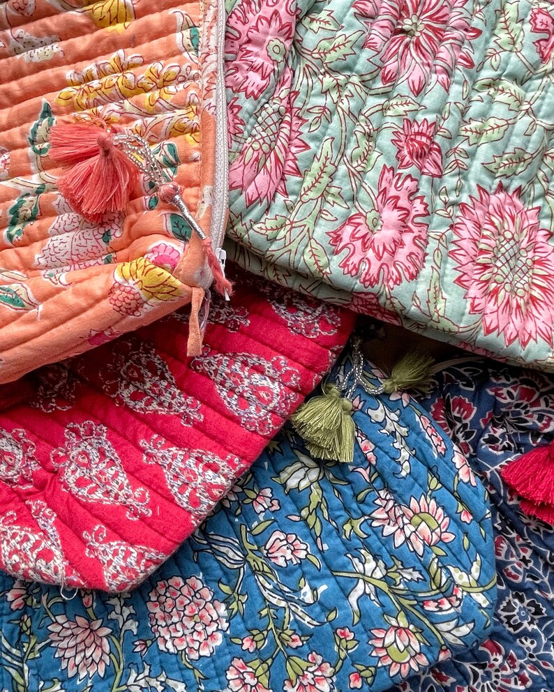 May include: Close-up of several quilted cosmetic bags in various colors. The bags feature floral patterns in shades of pink, orange, blue, and red. Each bag has a tassel zipper pull.