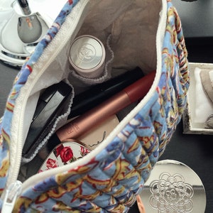 May include: Open blue floral quilted cosmetic bag with a white zipper, filled with makeup items. Includes a compact mirror with a floral design, a Chanel compact, and various tubes and applicators.