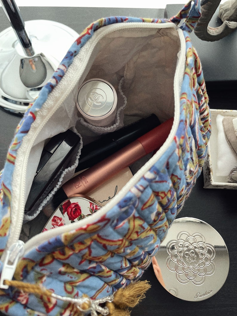 May include: Open blue floral quilted cosmetic bag with a white zipper, filled with makeup items. Includes a compact mirror with a floral design, a Chanel compact, and various tubes and applicators.