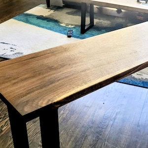 Peut inclure: Une console rectangulaire dotée d'un plateau en bois naturel avec un détail de bord brut. La table est soutenue par deux pieds en métal noir en forme de U qui assurent une base stable. La surface en bois présente un ton brun chaud avec des veines.