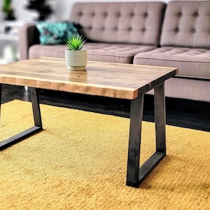 May include: A wooden coffee table with a live edge top and black metal legs. The table is sitting on a yellow rug in front of a gray sofa.