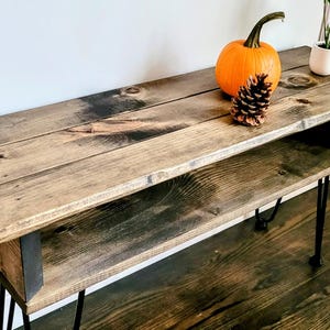 May include: A rustic wooden console table with a shelf, stained in a dark brown color. The table has black hairpin legs and is decorated with a small orange pumpkin and a pine cone.