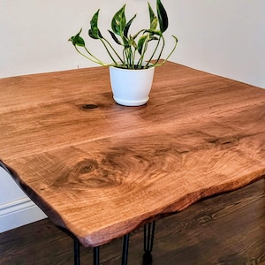 May include: A square wooden table with a natural live edge finish and a rich brown grain pattern. The table is supported by four thin black metal hairpin legs. A small green plant in a white pot sits in the center of the tabletop surface.